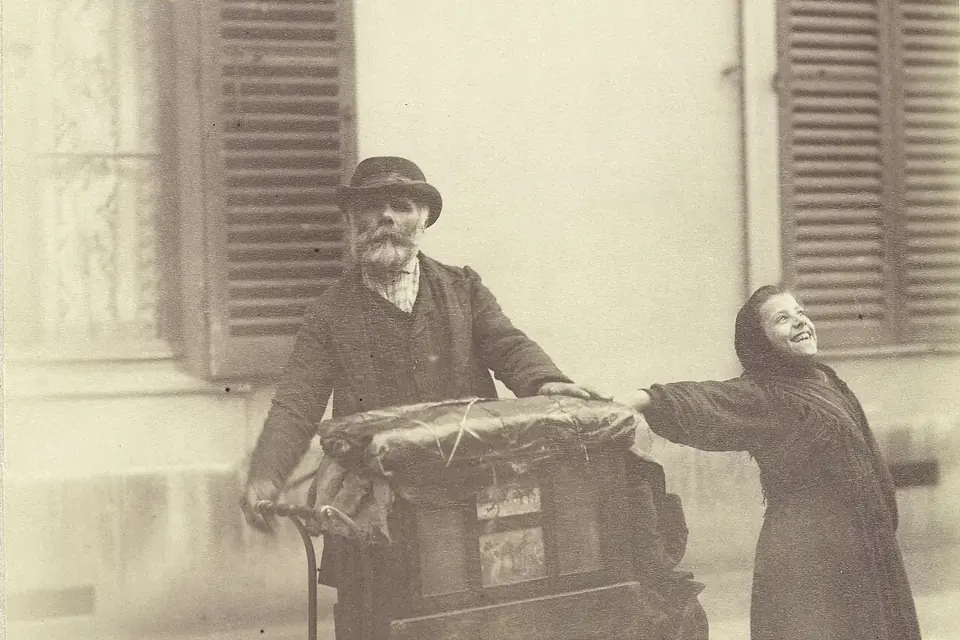 Photographie ancienne sépia d'un joueur d'orgue de Barbarie en chapeau et d'une jeune chanteuse des rues devant une fenêtre à volets