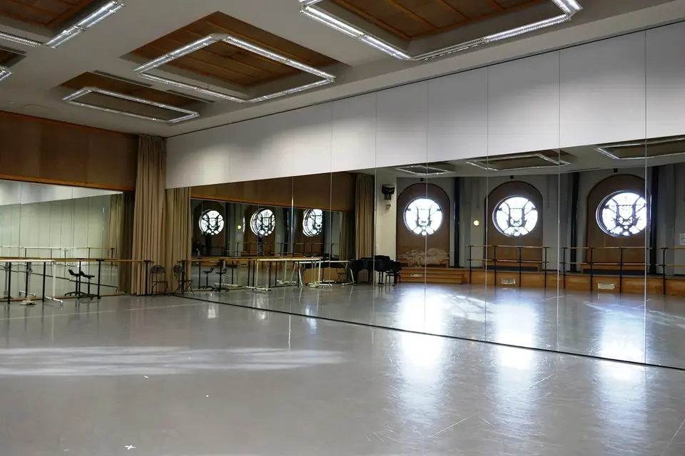 Grande salle de répétition vide au parquet clair, avec miroirs, barres et fenêtres rondes sous un plafond à caissons